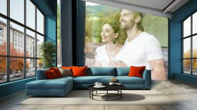 Young and lovely couple standing together looking outside the window at home, view through the window vith reflection Wall mural