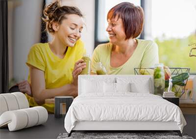 Young and older women sitting with healthy food and fresh drinks after the sports training indoors on the window background Wall mural