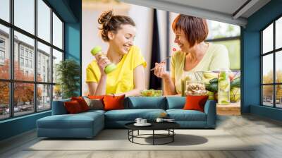 Young and older women sitting with healthy food and fresh drinks after the sports training indoors on the window background Wall mural