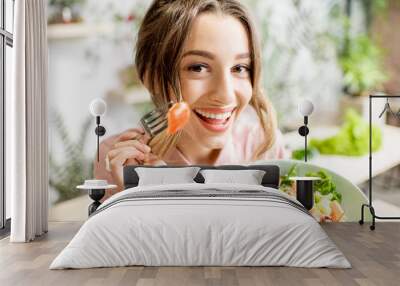 Young and playful woman eating healthy food sitting indoors on the green background Wall mural