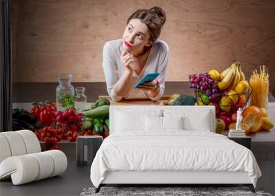 Young and pretty woman using smart phone sitting at the table full of fruits and vegetables in the wooden interior. Counting calories with mobile app. Food and health care concept Wall mural