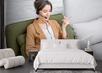 Young business woman working on computer, talking online using a headset while sitting on the comfortable sofa at home. Concept of remote work from home Wall mural