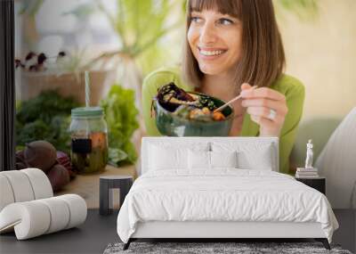 Young cheerful woman eats vegetarian lunch in bowl, sitting by the table full of fresh food ingredients indoors. Healthy lifestyle and wellness concept Wall mural