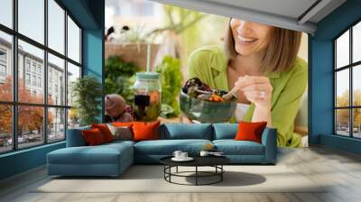 Young cheerful woman eats vegetarian lunch in bowl, sitting by the table full of fresh food ingredients indoors. Healthy lifestyle and wellness concept Wall mural