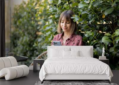 Young cheerful woman using smart phone while sitting on background of green bushes outdoors. Concept of online communication and spending time with gadgets outdoors Wall mural