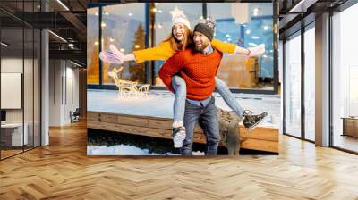 Young couple dressed in bright sweaters and hats having fun together near the decorated house during the winter holidays Wall mural