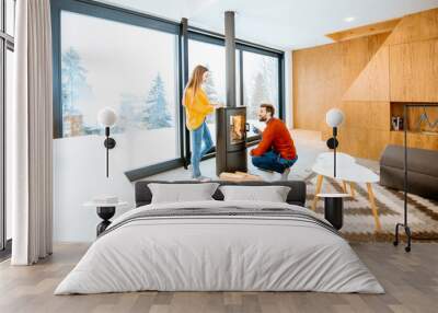 Young couple dressed in bright sweaters warming up near the fireplace in the modern house in the mountains during the winter Wall mural