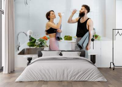 Young couple in sportswear having a break after the yoga training, drinking water at the kitchen with healthy food at home Wall mural