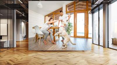 Young couple playing with dog during a breakfast in the dining room of their beautiful wooden country house Wall mural