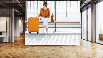 Young female traveler sitting with a suitcase at the transport stop, waiting for the tram or bus. Direct angle view on a stop bench. Concept of a city transportation and travel Wall mural