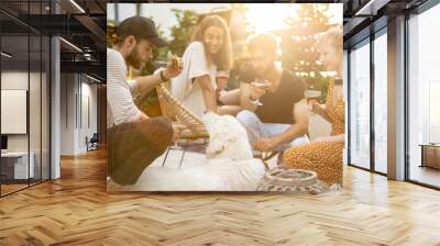 Young friends hanging out together, sitting with a dog, drinking wine and talking. People spending summer time having lunch together at backyard of country house at sunset Wall mural