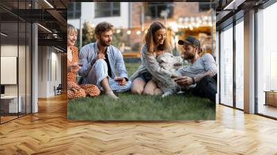 Young friends having fun playing with a dog and drinking wine, sitting on the green lawn at backyard of the country house in the evening Wall mural