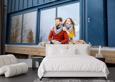 Young lovely couple dressed in colorful sweaters enjoying nature sitting together with hot drinks on the terrace of the modern house in the mountains Wall mural