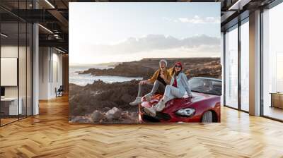 Young lovely couple enjoying landscapes, sitting together on a car hood, traveling by car on the rocky ocean coast. Carefree lifestyle, love and travel concept Wall mural