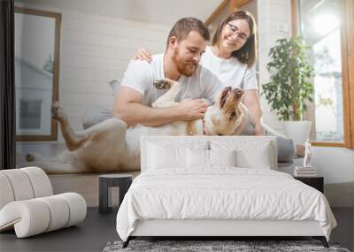 Young lovely couple in white t-shirts playing with their dog sitting on the floor in the house Wall mural