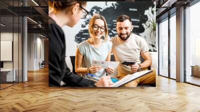 Young lovely couple sitting with manager at the travel agency office prepairing for a summer vacation Wall mural