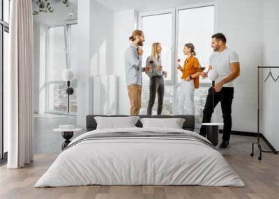 Young people dressed casually meeting in the white spacious hallway or showroom, talking and drinking wine during some informal event Wall mural