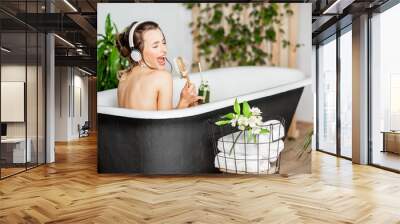 Young relaxed woman listening to the music and drinking smoothie while lying in the retro bathtub at the beautiful bathroom with green plants Wall mural