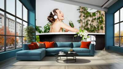 Young relaxed woman listening to the music and drinking smoothie while lying in the retro bathtub at the beautiful bathroom with green plants Wall mural