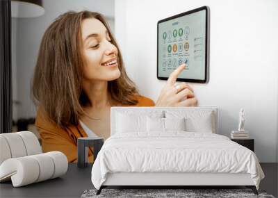 Young woman controlling home with a digital touch screen panel installed on the wall in the living room. Concept of a smart home and mobile application for managing smart devices at home Wall mural