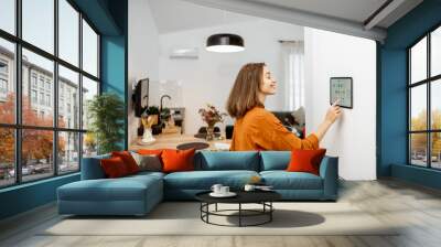 Young woman controlling home with a digital touch screen panel installed on the wall in the living room. Concept of a smart home and mobile application for managing smart devices at home Wall mural