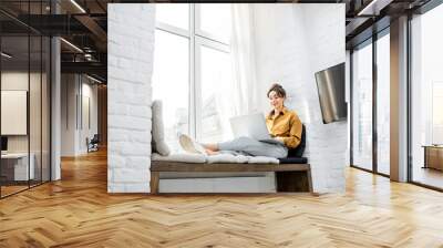 young woman dressed casually working on laptop while sitting on the window sill at home. work from h Wall mural