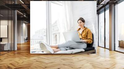 Young woman dressed casually working on laptop while sitting on the window sill at home. Work from home at cozy atmosphere concept Wall mural