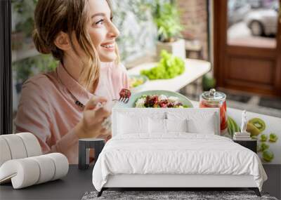 Young woman eating healthy food sitting in the beautiful interior with green flowers on the background Wall mural