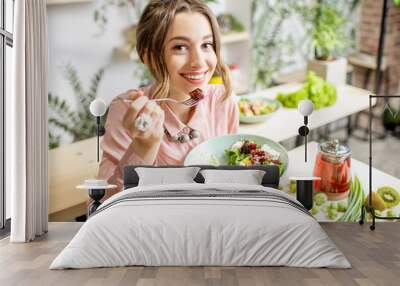 Young woman eating healthy food sitting in the beautiful interior with green flowers on the background Wall mural