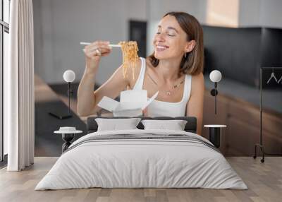 Young woman eating noodles with chopsticks from cardboard packaging in the kitchen at home. Takeaway asian food, ordering online concept Wall mural