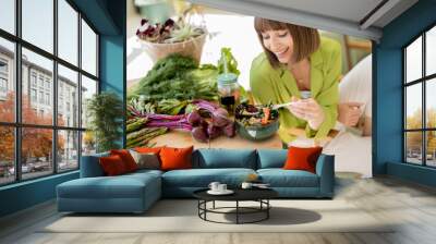 Young woman eats vegetarian lunch in bowl while sitting on chair near table with lots of fresh food ingredients in room with green plants. Healthy lifestyle concept Wall mural