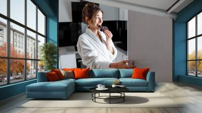 Young woman in bathrobe eating chocolate near the fridge, feeling hungry at night. Concept of not regular eating and overeating at night Wall mural