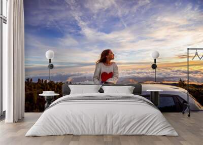 young woman in sweater with heart shape enjoying beautiful cloudscape sitting on the car roof above  Wall mural