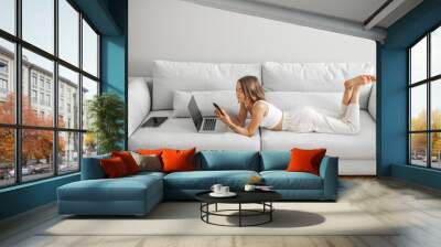 Young woman lying relaxed with phone and laptop on the comfortable couch at home. Front view. Working or leisure time at home Wall mural