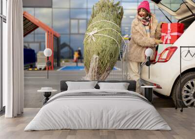 Young woman packing Christmas tree and presents into car trunk at parking lot of a mall. Cheerful girl having festive shopping in anticipation of the winter holidays Wall mural