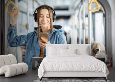 Young woman passenger standing with headphones and smartphone while moving in the modern tram, enjoying trip at the public transport Wall mural