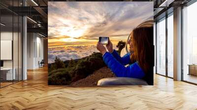 Young woman photographing with smart phone beautiful cloudscape looking out the car window on the roadside above the clouds on the sunrise Wall mural