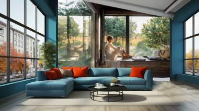 Young woman resting at beautiful country house or hotel, sitting on the window sill enjoying beautiful view on pine forest. Concept of solitude and recreation on nature Wall mural