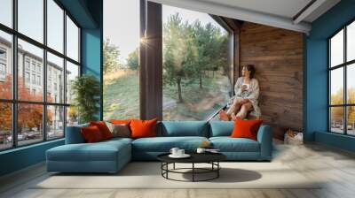 Young woman resting at beautiful country house or hotel, sitting with phone and cup on the window sill enjoying beautiful view on pine forest. Concept of solitude and recreation on nature Wall mural