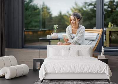 Young woman resting on sunbed and looks on the phone on terrace at modern house with panoramic windows near pine forest. Concept of solitude and recreation on nature. Wellness and mindful resort. Wall mural