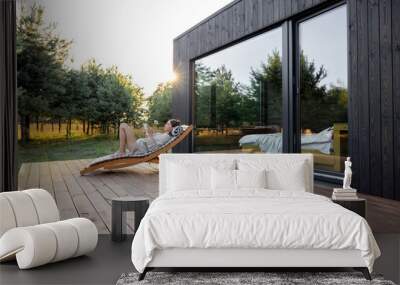 Young woman resting on sunbed and reading on a tablet on the wooden terrace near the modern house with panoramic windows near pine forest. Concept of solitude and recreation on nature Wall mural