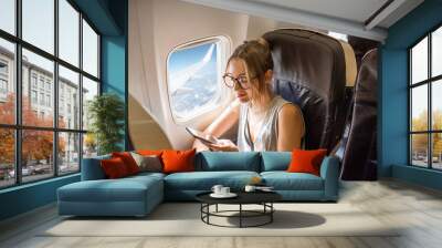 Young woman sitting with phone on the aircraft seat near the window during the flight in the airplane Wall mural