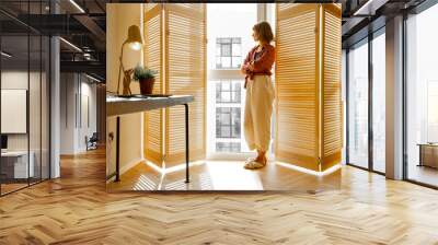 Young woman stands alone by the window blinds at cozy and sunny living room of modern apartment in beige tones. Quarantine, loneliness and life at home Wall mural