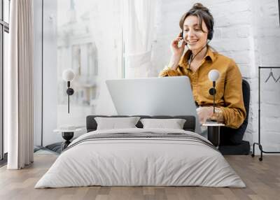 Young woman with a headset working online on computer while sitting on the window sill at home. Concept of studying or working from home online Wall mural