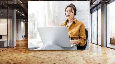 Young woman with a headset working online on computer while sitting on the window sill at home. Concept of studying or working from home online Wall mural