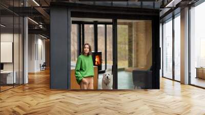 Young woman with her dog at modern house on nature. Girl in green sweater standing near the window and look outside. Concept of comfort and happy modern life Wall mural