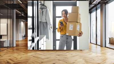 Young woman with parcels at the hallway at home, holds smart phone and checking goods. Concept of buying online and delivering goods home Wall mural