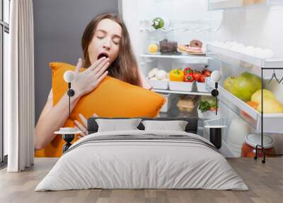 Young woman with pillow yawning standing in front of the refrigerator. Concept of eating during the night Wall mural