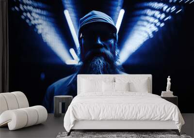 A man with beard, wearing a black hat facing away from light beams, blue neon light background Wall mural