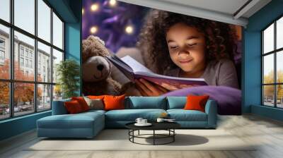 Little child kid is reading the storybook in evening in dark, sleeping in the bed, hugging her toy bear doll. Wall mural